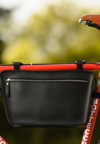 Svart läder cykelväska med en ficka fram med dragkedja, rektangulär form, fäst vid en röd cykelram, med synlig sömnad och metallaccent.