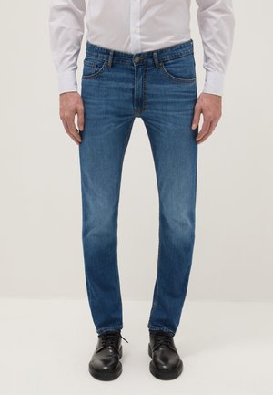 Homme portant un jean bleu slim, une chemise blanche boutonnée et rentrée, et des chaussures habillées en cuir noire, debout les mains détendues le long du corps.