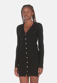 Abito nero a coste con maniche lunghe, scollatura a V e dettagli con bottoni argentati lungo il davanti. Design aderente con orlo a metà coscia.