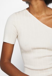 Jasno beżowy, prążkowany sweter z krótkim rękawem i asymetrycznym dekoltem. Teksturowany materiał podkreśla pionowe linie.