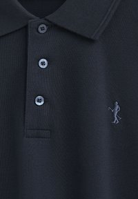 Svart polo skjorta i texturerad tyg med tre knappar och en liten broderad figur på vänster bröst.