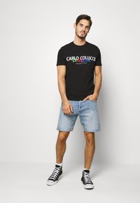 Sort T-shirt med farverig tekst, der læser "CARLO COLUCCI MILANO ITALY," parret med lyse denimshorts og sorte sneakers.