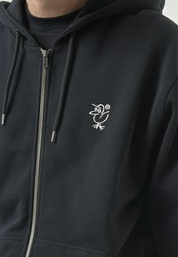 Schwarze Zip-Hoodie aus Baumwollmischung mit einem weißen gestickten Vogel-Logo auf der linken Brustseite und einer Kapuze mit Kordelzug.