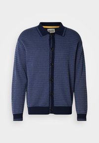 Μπλε ναυτικό πλεκτό ζακέτα με υφαντό σχέδιο, γιακά, κουμπιά μπροστά και αντιθετικές λεπτομέρειες κατά μήκος της πατιλέτας και των μανσετών.