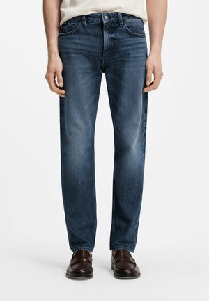 Mann steht, trägt ausgewaschene dunkelblaue Jeans mit geradem Bein, weißes Hemd und dunkelbraune Lederloafers vor weißem Hintergrund.