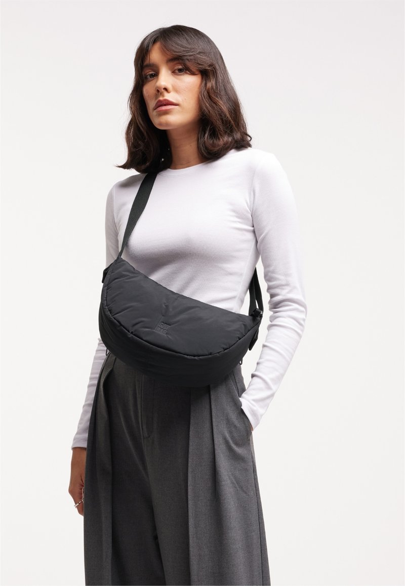 Sac en bandoulière rembourré noir, de forme courbée, avec une sangle ajustable et un logo discret ; porté sur un haut à manches longues blanc.