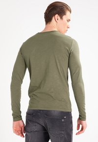 Long-sleeve olive green shirt with a round neckline and a smooth texture, worn under gray jeans, showing a minimalist design.