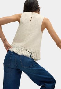 Top senza maniche intrecciato color avorio con orlo a frange e dettaglio a chiave sul retro, abbinato a jeans a gamba larga blu scuro.