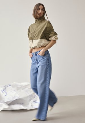 Frau in einer kurzen olivgrünen und beigen Jacke und lockeren blauen Jeans, die mit den Händen in den Taschen vor einem einfarbigen hellen Hintergrund steht.