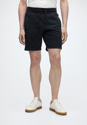Mand iført sorte shorts med snøre, hvide sneakers med gummisåler og en hvid skjorte, stående mod en ensfarvet lys baggrund.