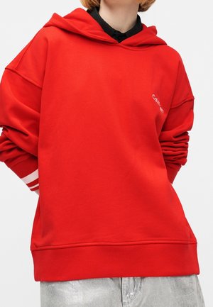Personne portant un sweat à capuche rouge vif à manches longues avec un petit logo blanc sur la poitrine, associé à un pantalon gris clair.