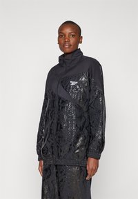 Black track jacket with shiny snake pattern and high collar. Features zipper front and Reebok logo on chest. Textured fabric with elastic cuffs.