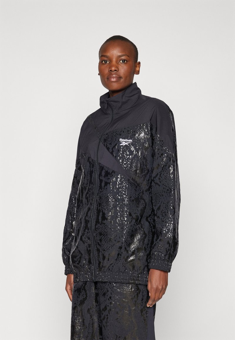 Black track jacket with shiny snake pattern and high collar. Features zipper front and Reebok logo on chest. Textured fabric with elastic cuffs.