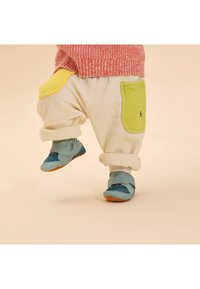 Pantalones crema de algodón con bolsillos acolchados en verde y amarillo, combinados con zapatos suaves en azul y turquesa que presentan un diseño de gato.