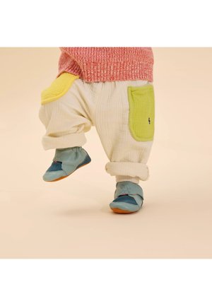 Baumwollcreme-Hosen mit grün- und gelb-gepolsterten Taschen, kombiniert mit weichen blauen und türkisfarbenen Schuhen mit einem Katzenmuster.