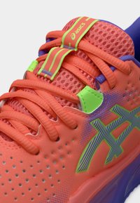 Φωτεινό πορτοκαλί αθλητικό παπούτσι με πλέγμα στην επιφάνεια, πράσινες και μωβ λεπτομέρειες, υφή στη σόλα και έντονο λογότυπο Asics στο πλάι.