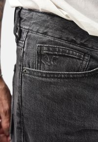 I jeans in denim nero presentano una tasca posteriore con dettagli di cucitura e un piccolo logo. Il tessuto ha un aspetto strutturato che ne evidenzia la durata.
