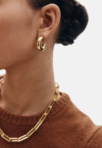 Pendiente de aro escultórico de oro con un acabado liso y brillante. Se lleva junto a un collar de cadena dorada enlazada y un suéter marrón.