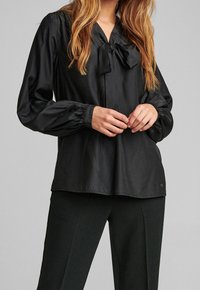 Blouse en satin noir avec un col en V et un détail nœud papillon, dotée de manches longues bouffantes et d'une texture lisse. Agrémentée d'un petit logo sur l'ourlet.