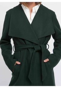 Dunkelgrüner Wollmantel mit weitem Kragen, langen Ärmeln, tailliertem Gürtel und seitlichen Taschen. Der Stoff wirkt weich mit einer strukturierten Oberfläche.