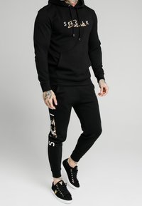 Set di felpa nera con cappuccio e pantaloni della tuta con accenti dorati. Tessuto morbido, vestibilità comoda, dotato di tasca a marsupio e polsini a caviglia affusolati. Sneakers nere.
