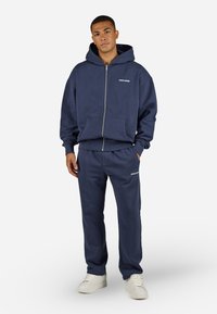 Navy blue zip-up hoodie with a front pocket, paired with matching sweatpants featuring an embroidered logo. Smooth texture, casual fit.