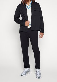 Svart långärmad skjorta över en krämfärgad kabelstickad tröja med blå detaljer, i kombination med svarta byxor och grå- och vita sneakers.