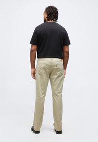 Homme aux cheveux tressés portant un t-shirt noir, un pantalon beige et des chaussures noires, debout face à un fond blanc, de dos à la caméra.