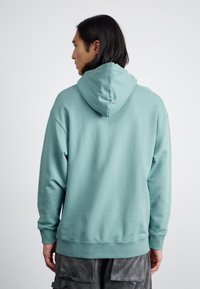 Felpa verde menta con cappuccio, polsini e orlo a coste. Tessuto morbido, vista da dietro, abbinata a pantaloni cargo grigi.