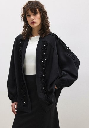 Nő fekete, túlméretezett kabátban, rajta virágmintás díszítésekkel, fehér felső és fekete szoknya fölött, sima világos háttér előtt áll.