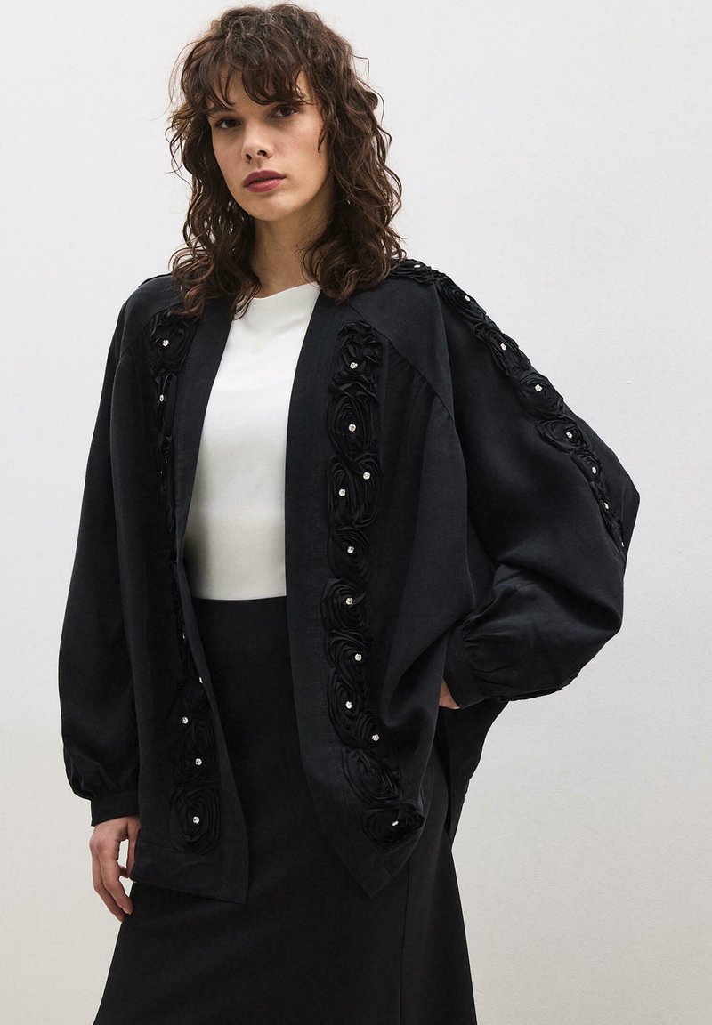 Γυναίκα που φοράει μαύρο φαρδύ μπουφάν με λουλουδάτες διακοσμήσεις πάνω από λευκή μπλούζα και μαύρη φούστα, στέκεται μπροστά σε απλό ανοιχτόχρωμο φόντο.