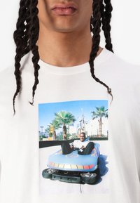 Tricou alb cu un grafic colorat al unei mașini de distracție albastre, cu o persoană zâmbitoare și palmieri în fundal.