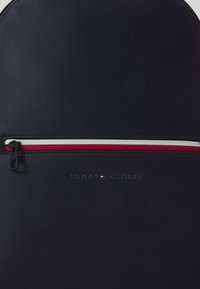 Zaino in finta pelle blu navy con una tasca frontale con zip e dettagli rossi e bianchi. Logo in rilievo nella parte inferiore, texture opaca complessiva.