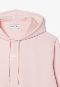 Světle růžová mikina s kapucí, klasického střihu, s regulovatelnou šňůrkou u kapuce, vyrobená z bavlny, s bílým logem Lacoste na přední straně.