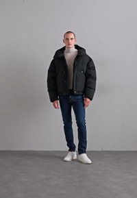 Mann steht gegen eine graue Wand und trägt eine schwarze Daunenjacke, einen beigen Rollkragenpullover, blaue Jeans und weiße Sneakers auf einem grauen Boden.