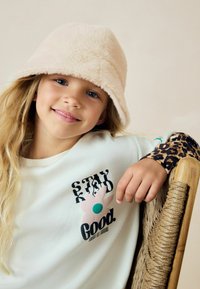Puszysty beżowy kapelusz typu bucket, biały T-shirt z kolorową grafiką kwiatu oraz akcenty w panterkę na rękawach. Ręka oparta na plecionej krześle.