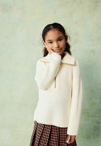 Pull en maille côtelée de couleur crème avec un col zippé à mi-hauteur, porté sur une jupe en tartan brun. La texture semble douce et tissée.