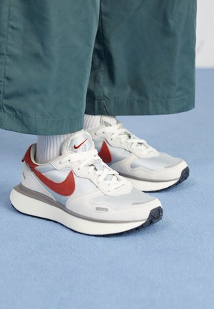 Sneakers Nike bianche e grigie con swoosh rosso, indossate con calzini bianchi e pantaloni verdi a gamba larga su pavimento con moquette blu.