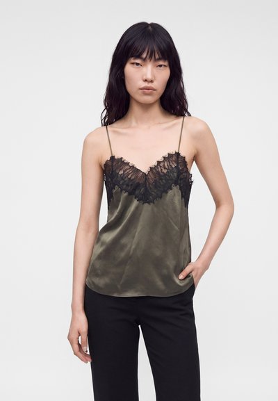 Femme aux longs cheveux noirs portant un caraco en satin vert olive avec une bordure en dentelle noire et fines bretelles, assorti à un pantalon noir, main dans la poche.