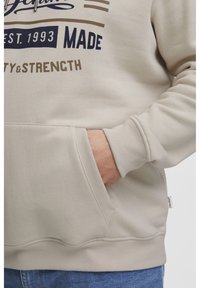 Sudadera de color beige claro hecha de tejido suave, con un bolsillo frontal, puños acanalados y texto impreso en marrón oscuro y claro.
