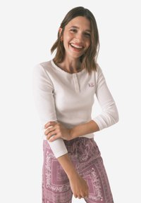 Camisetta bianca a maniche lunghe con scollatura rotonda e tre bottoni, dotata di ricami delicati. Abbinata a pantaloni rosa con motivo.