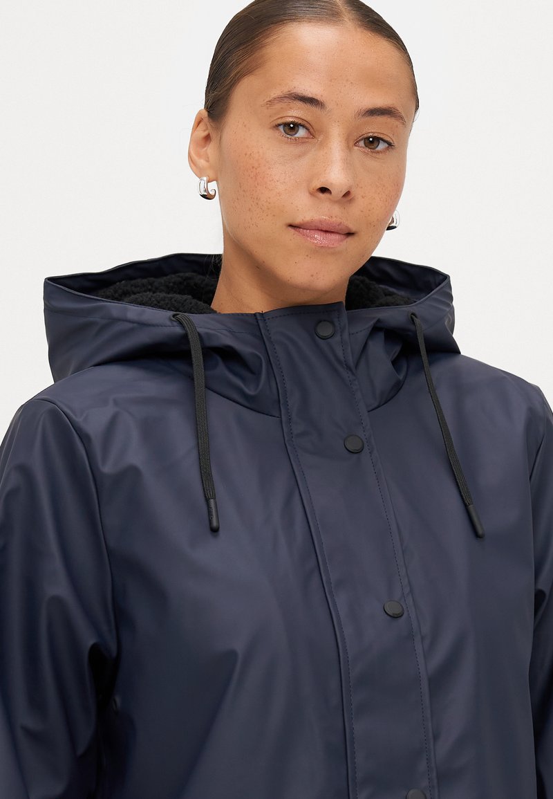 Veste imperméable navy avec capuche, pressions à l'avant et cordons de serrage. Doublure intérieure douce avec une finition texturée.