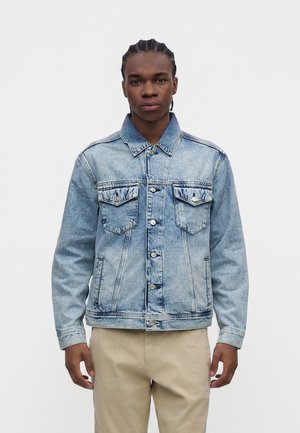 Junger Mann mit geflochtenem Haar trägt eine hellblaue Jeansjacke und beige Hose, steht vor einem schlichten weißen Hintergrund.