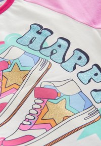 Balta krekla ar krāsainu sporta apavu grafiku un spīdīgu akcentu. "HAPPY" uzraksts spīdīgā zilā, rozā un daudzkrāsainā zvaigžņu rakstā.