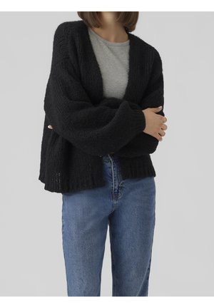 Personne portant un cardigan noir en laine ample sur une chemise grise, assorti à un jean bleu, les bras croisés devant un fond gris.