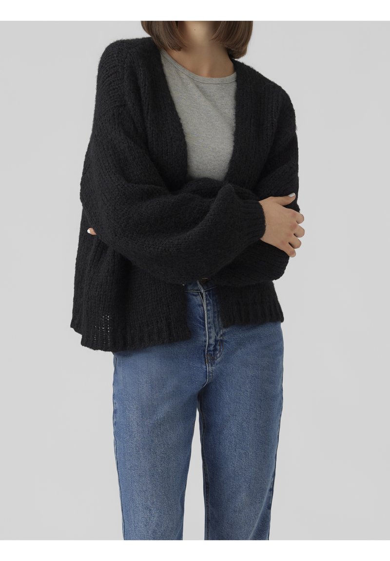 Personne portant un cardigan noir en laine ample sur une chemise grise, assorti à un jean bleu, les bras croisés devant un fond gris.