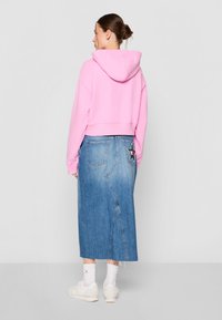 Felpa rosa tagliata con cappuccio e cordini, abbinata a una gonna maxi in denim blu con una toppa a forma di stella e uno spacco laterale. Sneakers bianche e calzini.