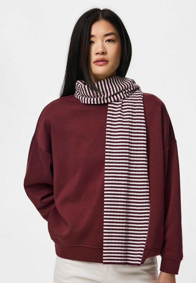 Femme aux longs cheveux noirs portant un pull marron et une écharpe rayée blanche et marron sur fond blanc.