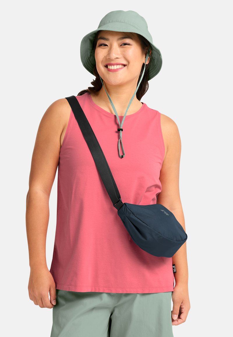 Femme souriante portant un chapeau bob vert, un haut sans manches rose, un short vert clair et un sac bandoulière noir sur un fond blanc.