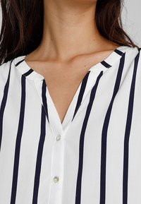 Blusa bianca con righe verticali blu navy, scollatura a V e bottoni sulla parte anteriore. Il tessuto è leggero e dalla superficie liscia.